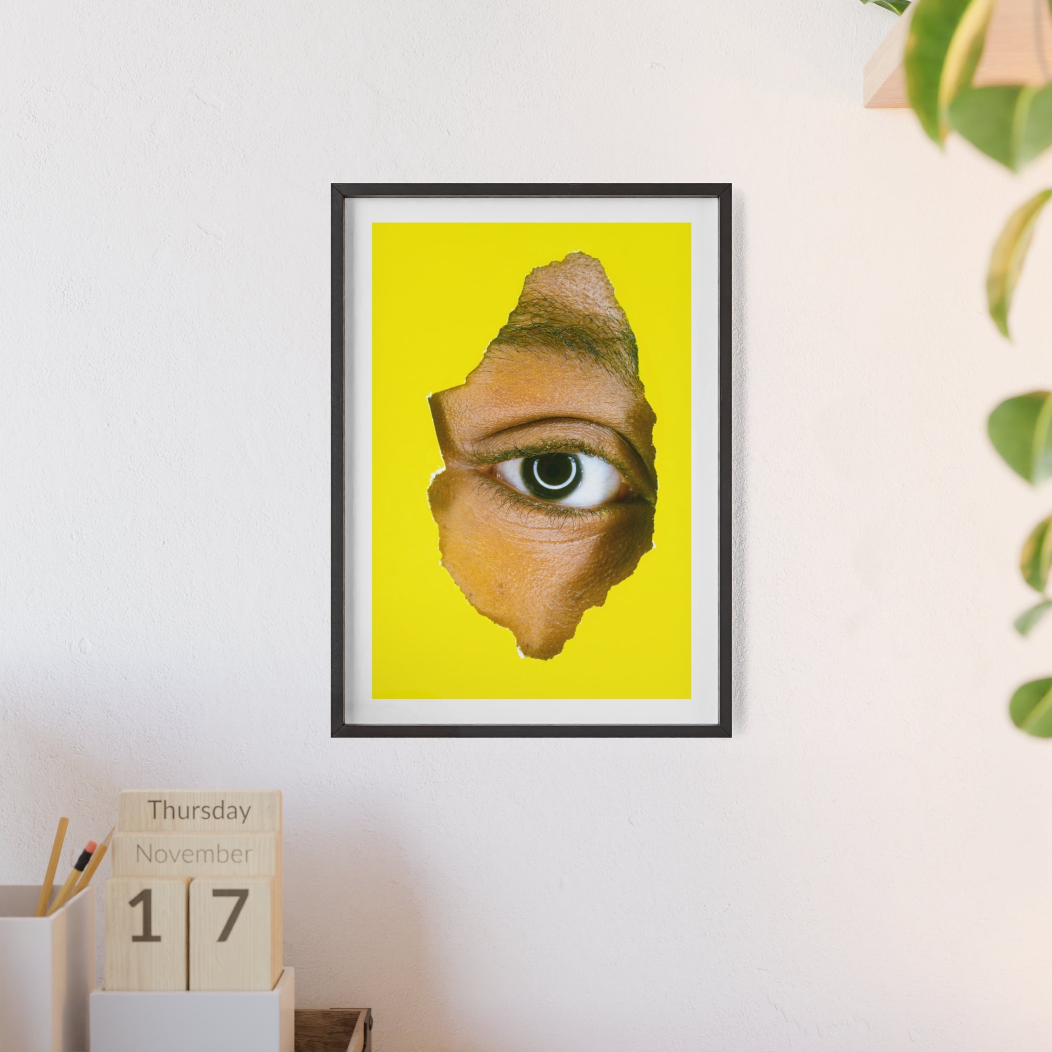 Framed artwork of an eye on a yellow background, displayed on a wall with a calendar and plant.