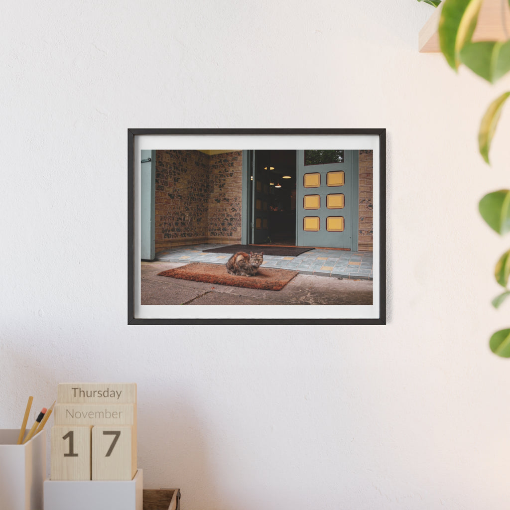 Framed photograph of a cat on a mat in front of a door, displayed on a wall with a calendar below.