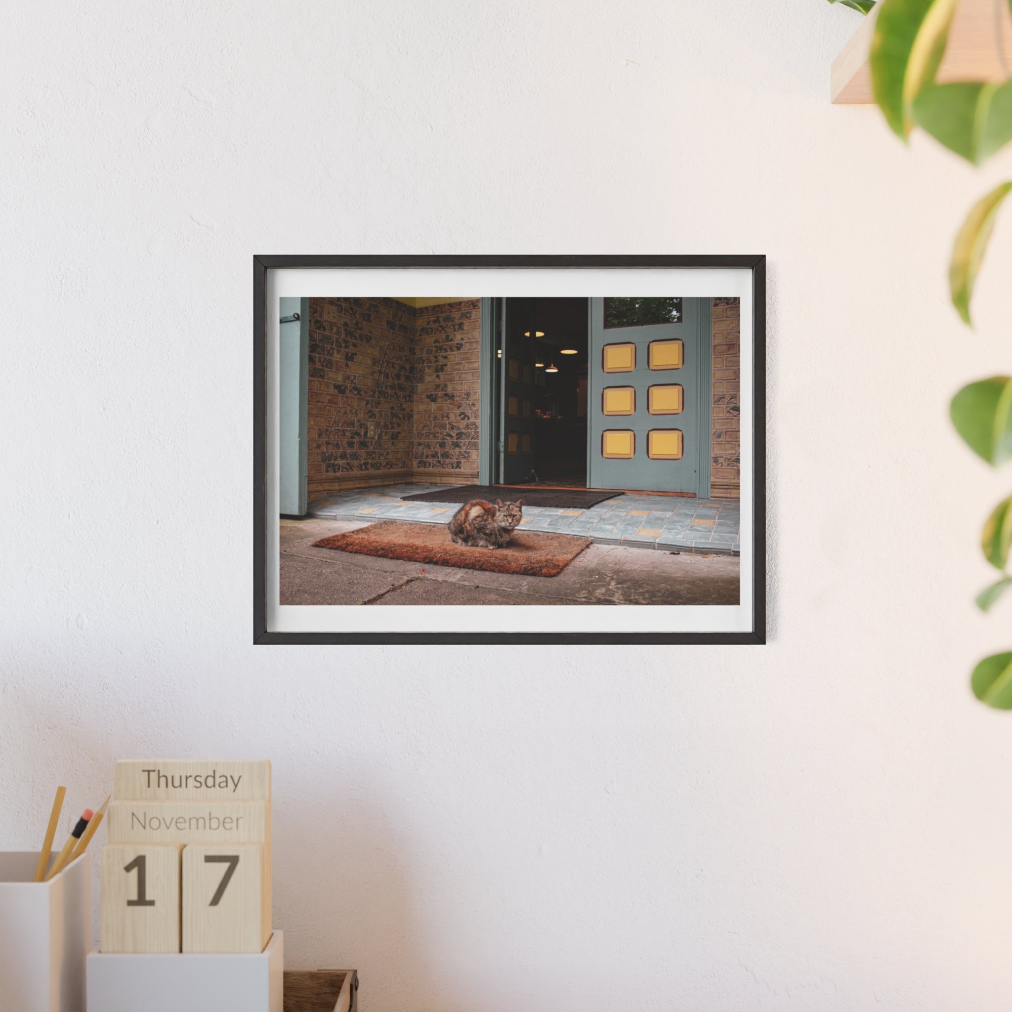 Framed photograph of a cat on a mat in front of a door, displayed on a wall with a calendar below.