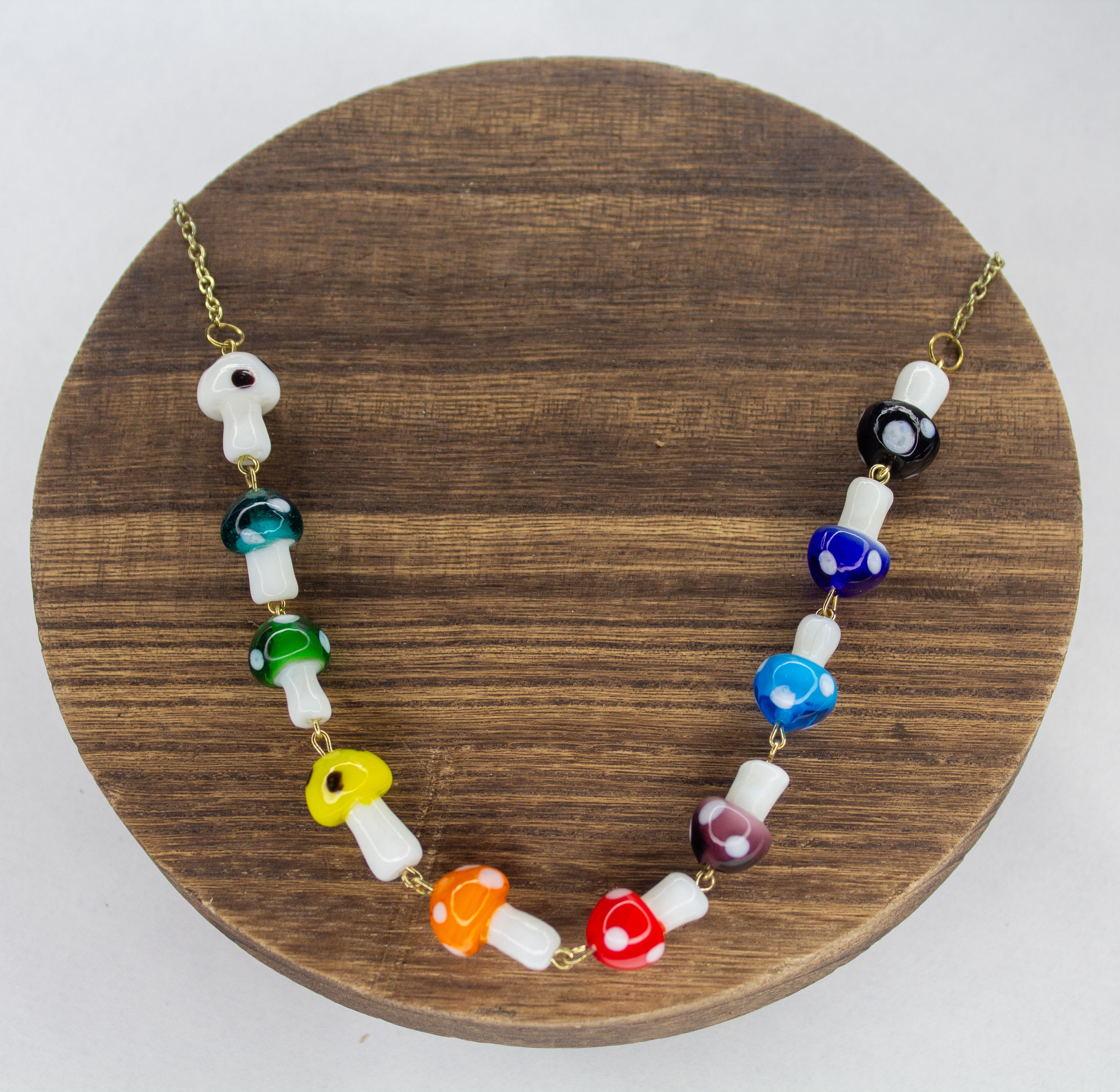 A necklace with multicolored glass mushroom charms on a gold plated chain, displayed on a wooden round stand.